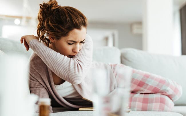Young woman coughing into elbow while lying down on sofa in the living room.