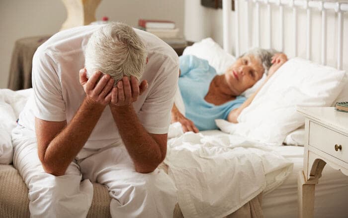 Worried Senior Man Sits Up On Bed Whilst Wife Sleeps.