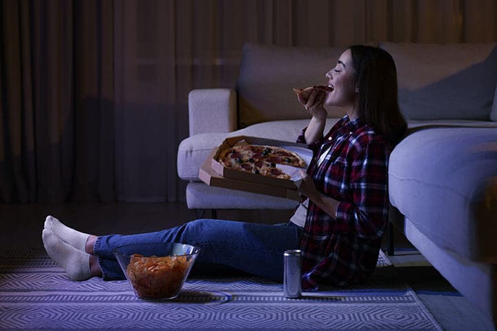 Young woman eating pizza while watching TV in room at night. Bad habit