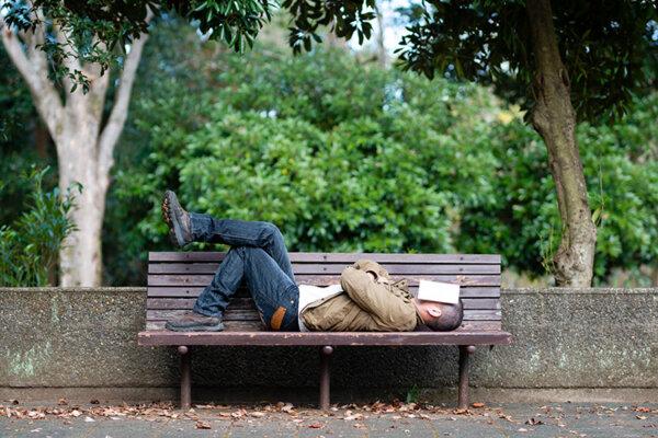 Mann schläft mit Buch im Gesicht auf einer Parkbank.