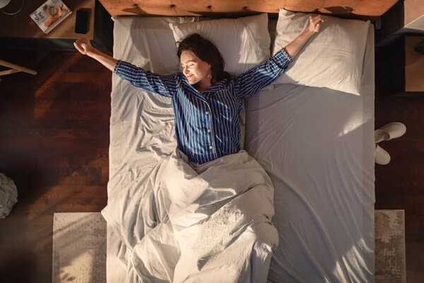 Junge Frau im Bett streckt sich in der durch das Fenster herein scheinenden Morgensonne nach erholsamem Schlaf.