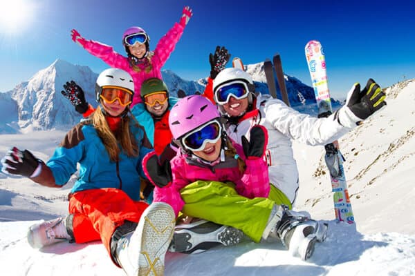 Fünfköpfige Familie mit Skiern und Skihelmen sitzt im Schnee und jubelt