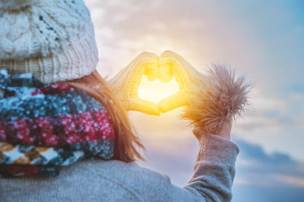 Eine Frau in einer Winterlandschaft formt mit den Händen ein Herz und umrahmt damit die untergehende Sonne.