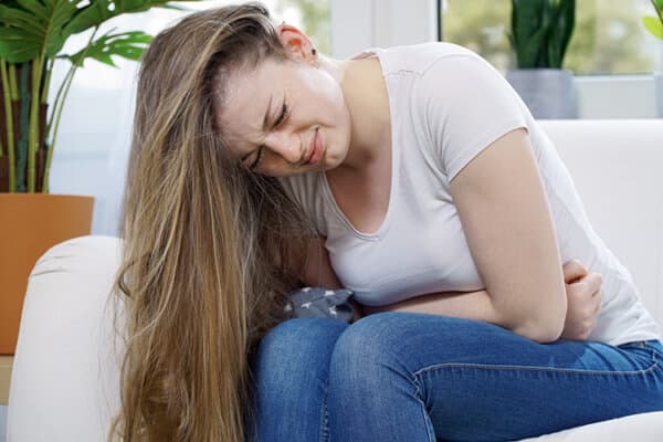 Junge Frau sitzt mit Menstruations-Schmerzen auf dem Sofa und hält sich mit schmerzverzerrtem Gesicht gekrümmt ihre Arme vor den Bauch.