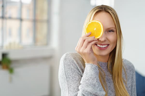 Eine junge blonde Frau sitzt in ihrem Wohnzimmer und hält sich lachend einen Schnitz Zitrone vor das Auge.